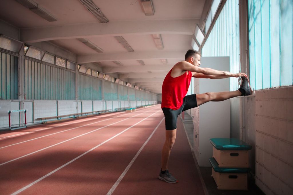 Coureur qui étire l’arrière de la cuisse, pied posé contre un mur de piste couverte