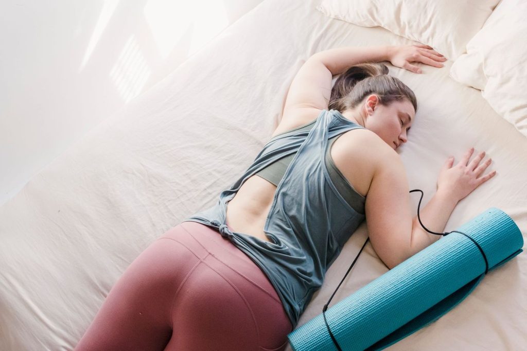 Femme en tenue de sport endormie sur un lit, tapis de yoga à côté